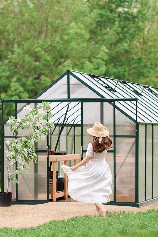 Woman in a white dress and straw hat standing in front of a greenhouse with greenery in the background