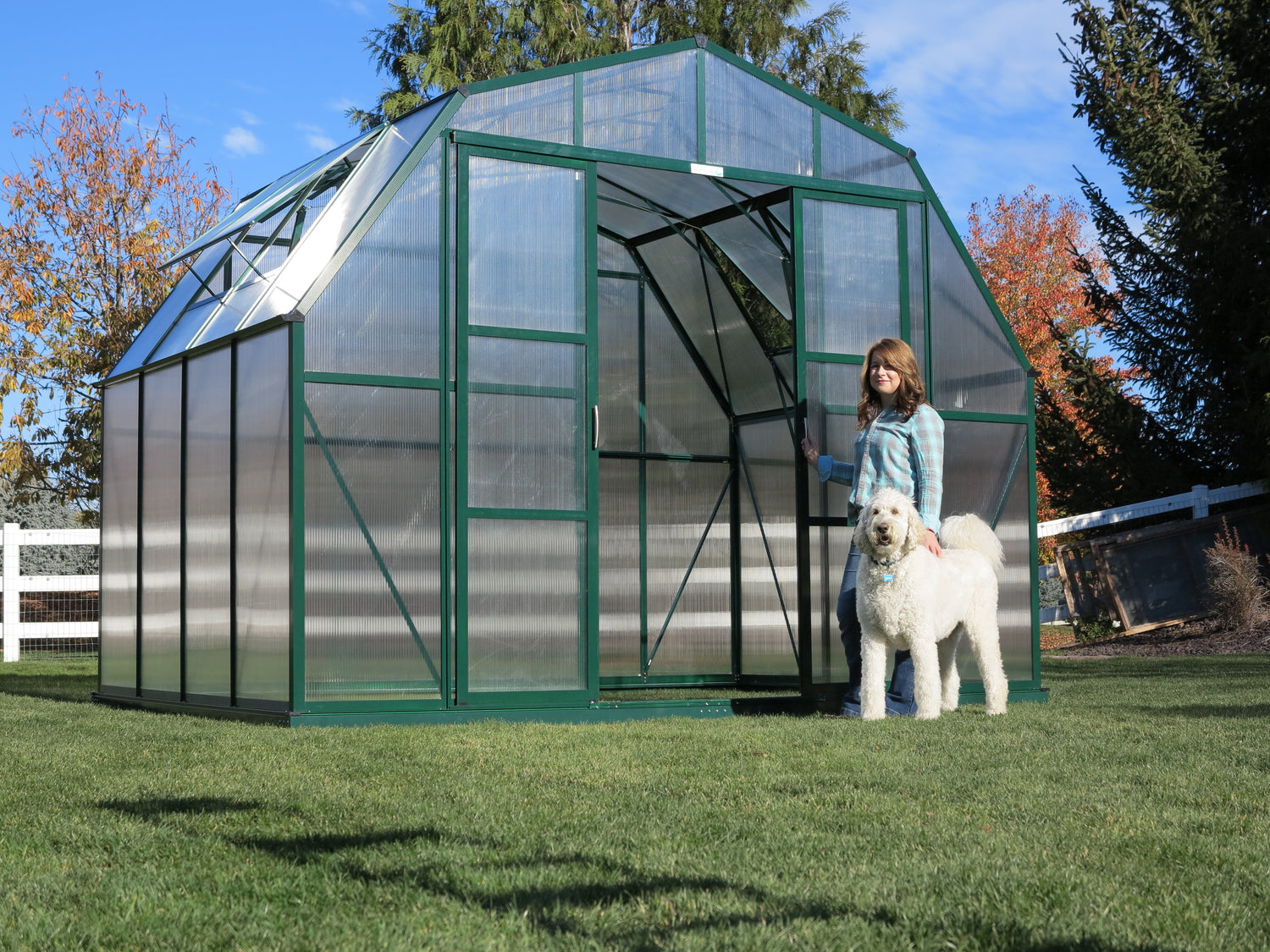 Person standing next to a large green greenhouse with a dog on a grassy area.